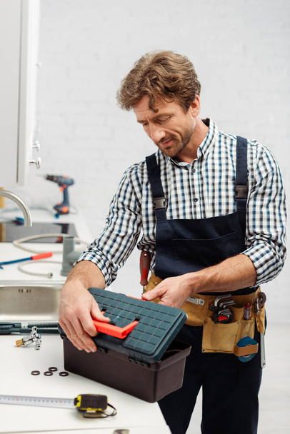 Выборочный фокус водопроводчика в коробке инструментов открытия рабочей одежды рядом с инструментами на столешнице на кухне
  - Фото, изображение