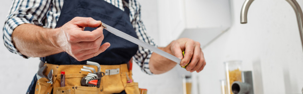 Panoramic shot of plumber holding industrial measuring tape in kitchen  - Фото, изображение