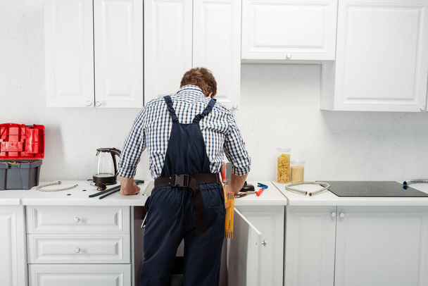 Vista trasera del fontanero en overol trabajando en cocina
  - Foto, Imagen