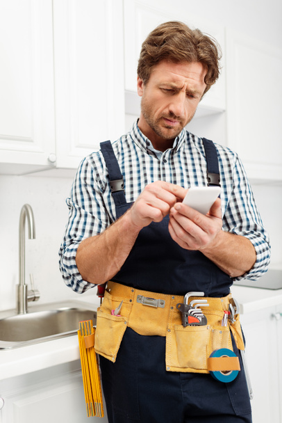 Selective focus of handsome plumber in tool belt and overalls using smartphone in kitchen  - Photo, Image
