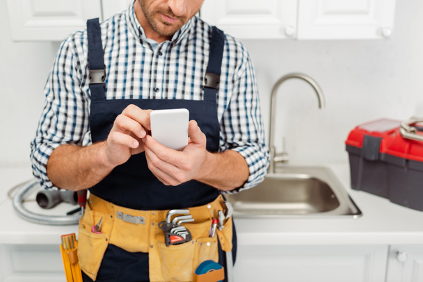 Vista recortada del trabajador en el cinturón de herramientas utilizando el teléfono inteligente mientras trabaja en la cocina
  - Foto, imagen
