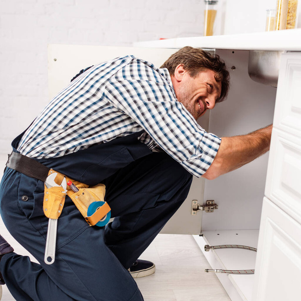 Side view of repairman in uniform and tool belt fixing kitchen sink  - Photo, Image
