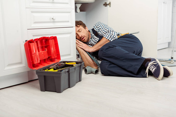 Klempner schläft auf dem Boden in der Nähe von Werkzeugkasten und Waschbecken in der Küche  - Foto, Bild