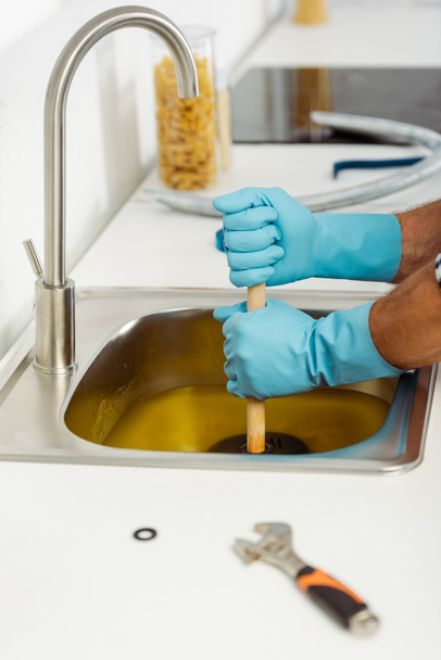 Vista recortada del trabajador en guantes de goma limpieza fregadero de cocina con émbolo cerca de la llave inglesa en la encimera
  - Foto, imagen