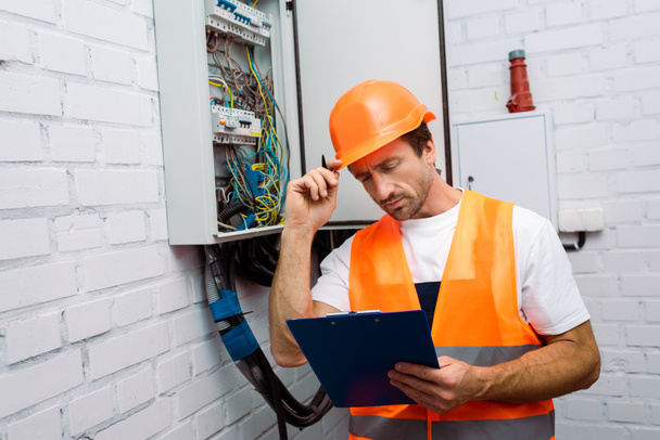 Guapo electricista sujetando portapapeles y bolígrafo mientras trabaja cerca del panel eléctrico
 - Foto, Imagen
