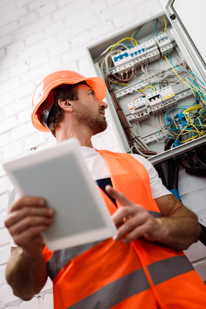 Selective focus of handsome electrician in hardhat looking at electrical distribution box while holding digital tablet  - Photo, Image
