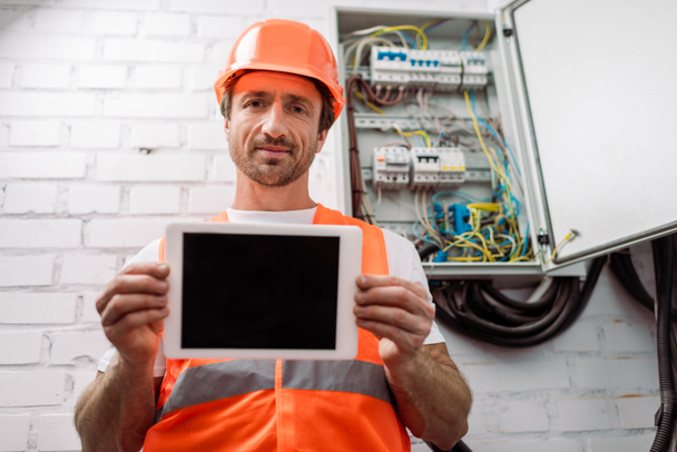 Selective focus of handsome electrician showing digital tablet with blank screen near electrical box - Photo, image