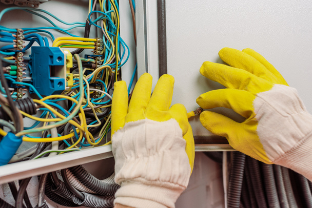 Oříznutý pohled na elektrikáře v rukavicích držící drát poblíž elektrického panelu - Fotografie, Obrázek