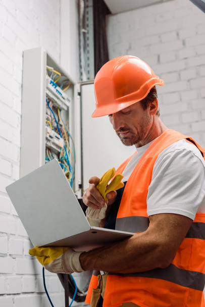 Handsome electrician pointing with finger while holding laptop near electrical box - Photo, Image