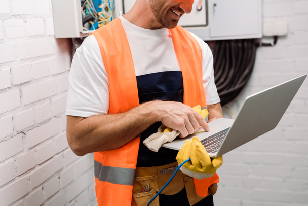 Oříznutý pohled na usmívajícího se elektrikáře držícího rukavici a používajícího notebook  - Fotografie, Obrázek