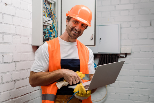 Smiling electrician in hardhat using laptop and looking at camera  - Foto, immagini