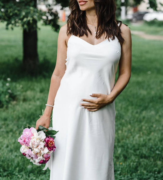 noiva grávida em um vestido fino branco em tiras finas com ombros nus com um buquê de peônias. maternidade
 - Foto, Imagem