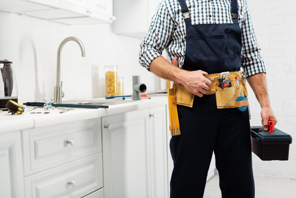 Gesneden weergave van loodgieter in werkkleding houden gereedschapskist in de buurt van gootsteen en kraan in de keuken  - Foto, afbeelding