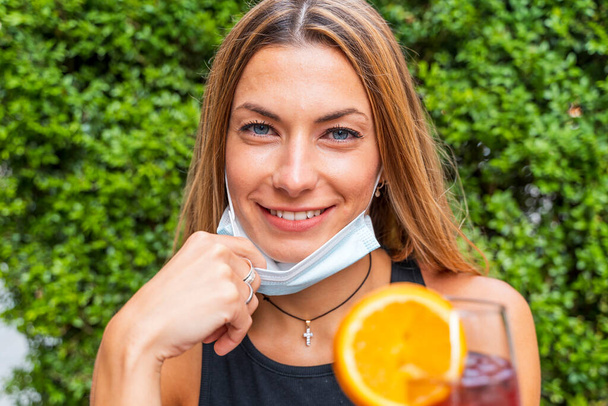female portrait of young adult drinking colored cocktail at outdoor bar with face masks - New normal lifestyles concept with happy people toasting drinks outside - Foto, Imagem