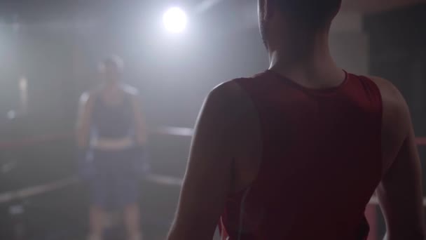 Caucasian boxers greeting each other on boxing ring. Young athletic sportsmen start sparring in dark foggy gym with backlight at the background. Sport, lifestyle, martial arts. - Footage, Video