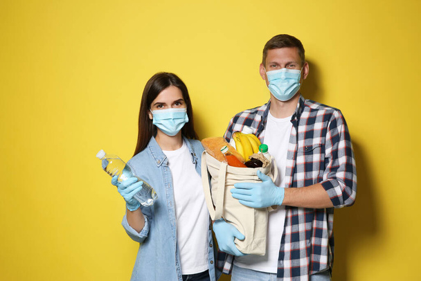 Volunteers in protective masks and gloves with products on yellow background. Aid during coronavirus quarantine - Fotografie, Obrázek