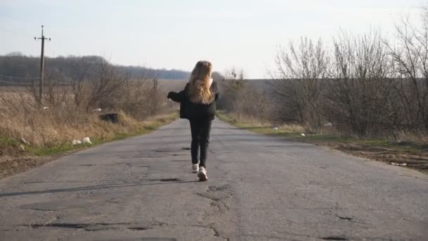 Rear view of little girl in jacket walks along empty road and dancing funny at sunny day. Small female child with long blonde hair haves fun on empty driveway at countryside. Slow motion - Footage, Video