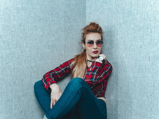 A beautiful girl in a red shirt with glasses and with a clock on her hand sits near the wall with gray wallpaper - Фото, изображение