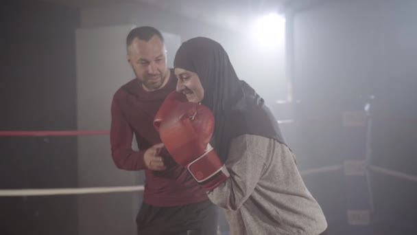 Positive muslim woman standing in boxing stance as Caucasian man talking at the background. Portrait of female boxer in hijab training with personal trainer in haze with backlight. - Video
