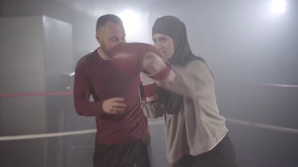 Coach correcting boxing stance of young beautiful muslim woman in hijab training in haze in backlight. Portrait of confident female boxer exercising with personal trainer in lense flare and fog. - Felvétel, videó