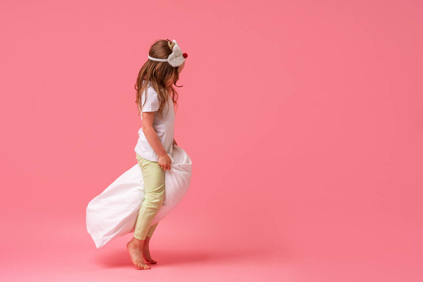 charming little girl in a sleep mask and pajamas jumps and flies on pillows and smiles on a pink background. The concept of healthy sleep. A copy of the space. In full growth. - Foto, Bild