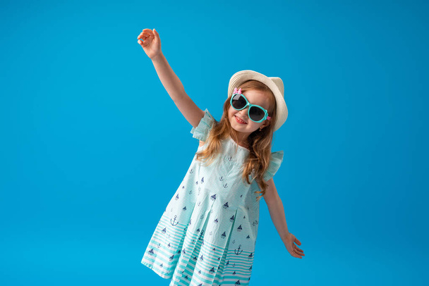 a cute little girl of 4 years old, in a dress, hat and sunglasses, poses, on a blue background. The child will be happy to start the summer holidays and go on a trip - Valokuva, kuva