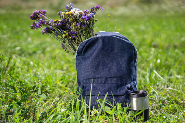 Χαλάρωση και καλοκαιρινή πεζοπορία έννοια. Μπλε τζιν σχολική τσάντα στέκεται στο πράσινο γρασίδι με ταξιδιωτική κούπα κοντά σε αυτό και όμορφα λουλούδια λιβάδι μέσα σε αυτό - Φωτογραφία, εικόνα