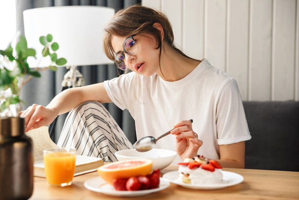 Photo of young focused woman reading book while having breakfast in cozy room at home - Foto, Imagem