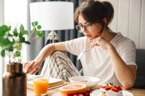 Photo of young focused woman reading book while having breakfast in cozy room at home - Photo, image