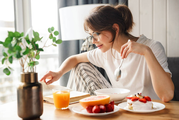 Photo of young pleased woman reading book while having breakfast in cozy room at home - Photo, image