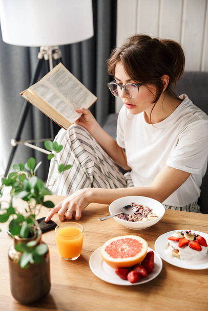 Photo of young woman reading book and using cellphone while having breakfast in cozy room at home - Foto, afbeelding