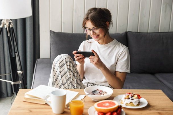 Photo of cheerful caucasian woman playing video game on cellphone while having breakfast in cozy room - Фото, зображення