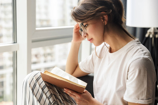 Photo of young thinking woman in eyeglasses reading book while sitting in cozy room at home - Фото, зображення