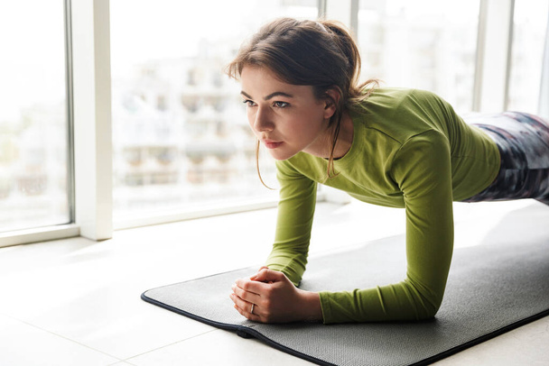 Photo of focused brunette woman doing yoga exercises on mat while working out in cozy room - Fotó, kép