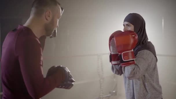 Side view of personal trainer training young confident muslim woman in hijab. Serious beautiful sportswoman listening to coach in foggy gym in backlight. Combat sport, gender equality, martial arts. - Video