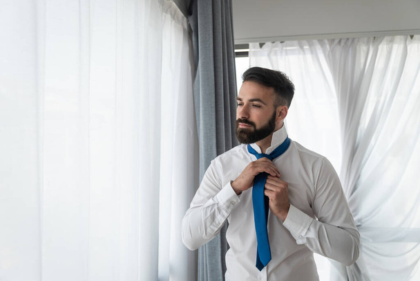 Jeune homme d'affaires barbu élégant élégant dans la chemise en essayant de nouer sa cravate au cou le matin avant qu'il ne se rende au travail
 - Photo, image