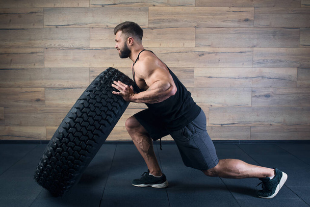 A strong man in a black tank top and grey shorts with a beard tries to push a tire in a gym. A muscular shredded guy doing a crossfit workout. A fellow with tattoos doing hardcore exercise.  - Photo, Image
