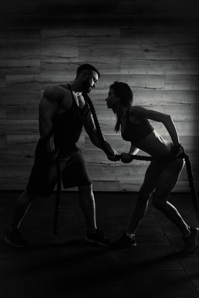 Egy sportos lány és egy szakállas, izmos férfi húzzák a kötelet a CrossFit edzés alatt egy edzőteremben. Egy testépítő edz egy fitt lánnyal. Sziluett. Fekete-fehér fénykép - Fotó, kép