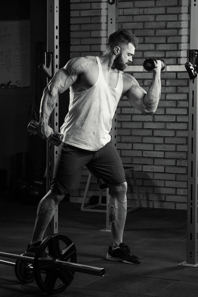 Un bodybuilder déchiqueté fait des boucles biceps avec des haltères. Un homme musclé avec des tatouages et une barbe dans un débardeur blanc et un short bleu s'entraîne dans une salle de gym. Une photo en noir et blanc. - Photo, image