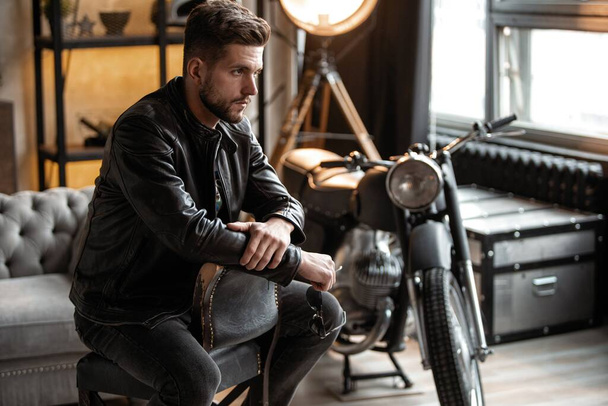 Handsome young man in leather jacket looking away while sitting on the stool - Foto, afbeelding