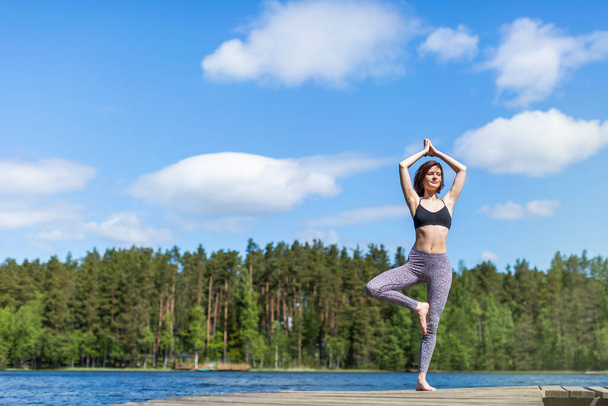 Woman practicing yoga on a lake standing on log in yoga tree pose. fitness lifestyle at the outdoors nature background. Sunny day. copy space  - Photo, image