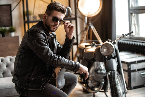Handsome young man in leather jacket looking away while sitting on the stool - Photo, image