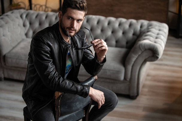 Handsome young man in leather jacket looking away while sitting on the stool - Fotografie, Obrázek