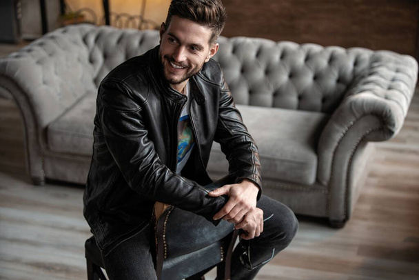 Handsome young man in leather jacket looking away while sitting on the stool - Fotoğraf, Görsel