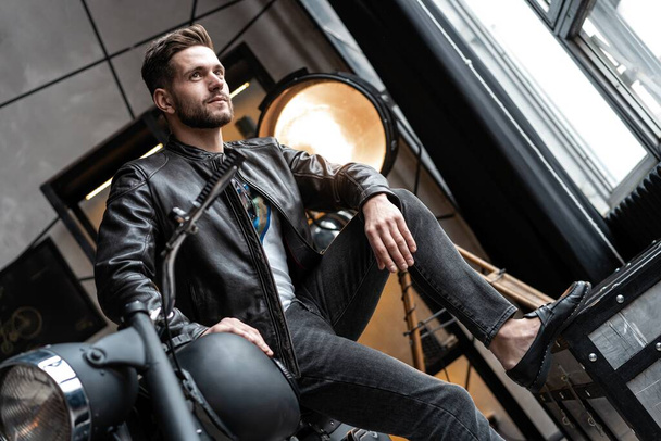 Handsome young man in leather jacket looking away while sitting on the stool - Foto, Imagen