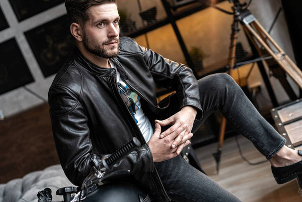 Handsome young man in leather jacket looking away while sitting on the stool - Φωτογραφία, εικόνα