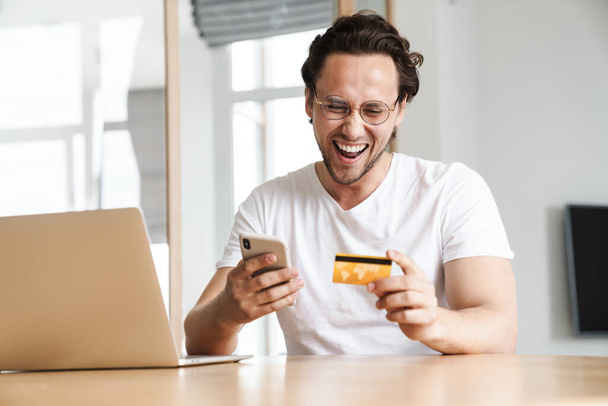 Attractive cheerful young man sitting at the kitchen table, working on laptop computer, shopping online with mobile phone and credit card - Foto, Bild