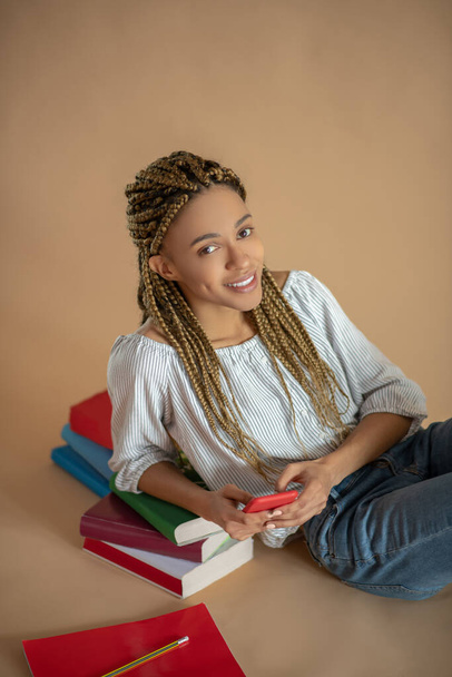 Young African American female sitting on the floor, leaning on pile of books, holding her mobile, smiling - Foto, immagini