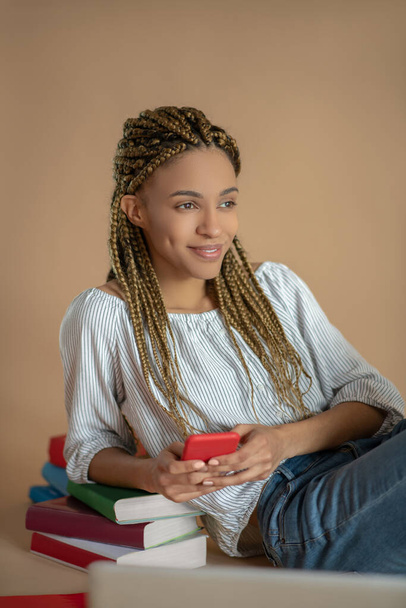 Young African American female sitting on the floor, leaning on pile of books, holding her mobile - Fotó, kép