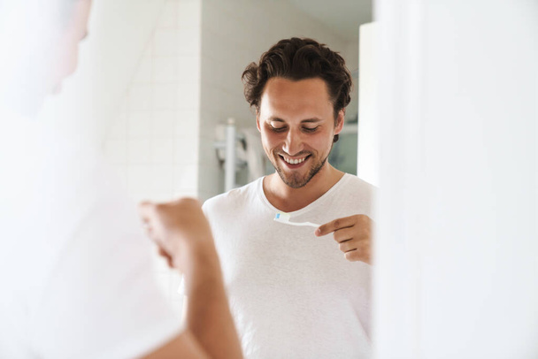 Attractive happy young man standing in front of the bathroom mirror, brushing teeth - Photo, Image